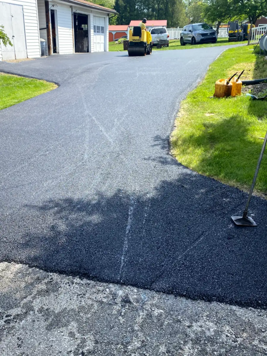 Freshly paved curving driveway beside a white garage with a roller working at the far end