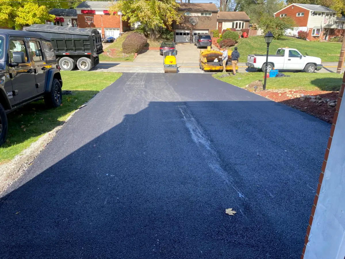 Fresh black driveway on a hillside street of brick split-level homes in fall, crew and roller at the top