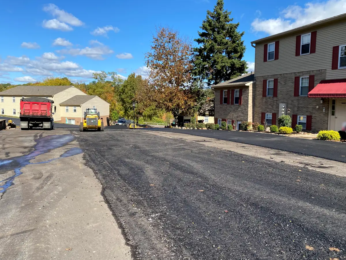 Crew paving a large apartment parking lot with a roller and dump truck under a blue sky