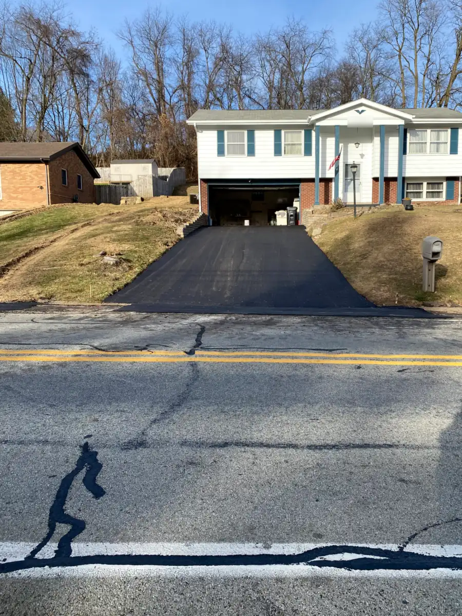 New asphalt driveway climbing to a split-level house with bare hardwoods on the hill behind