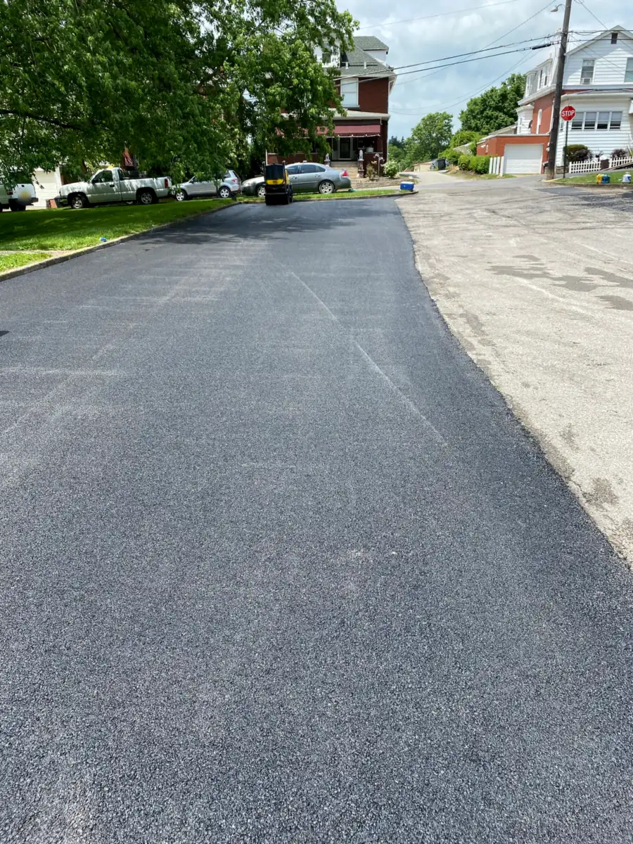 Long new asphalt drive beside a street of brick houses, with the roller finishing at the far end